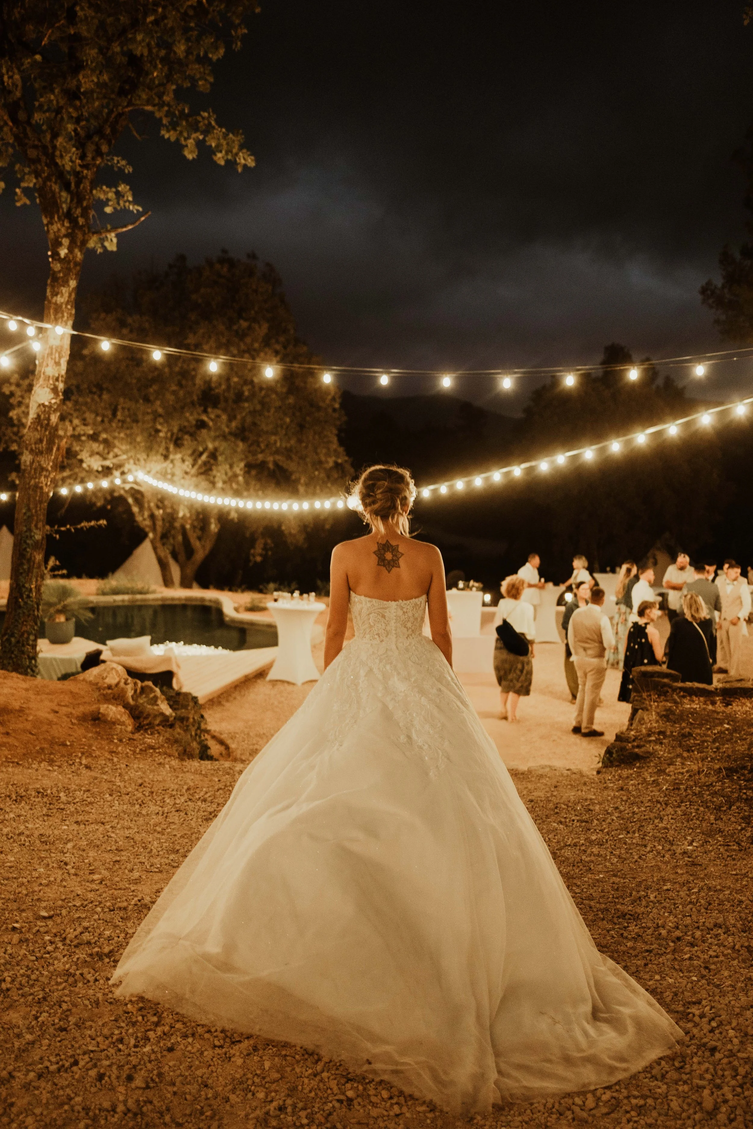 Une mariée en robe de mariage blanche vue de dos lors d'un mariage en extérieur la nuit, avec des invités et des guirlandes lumineuses.