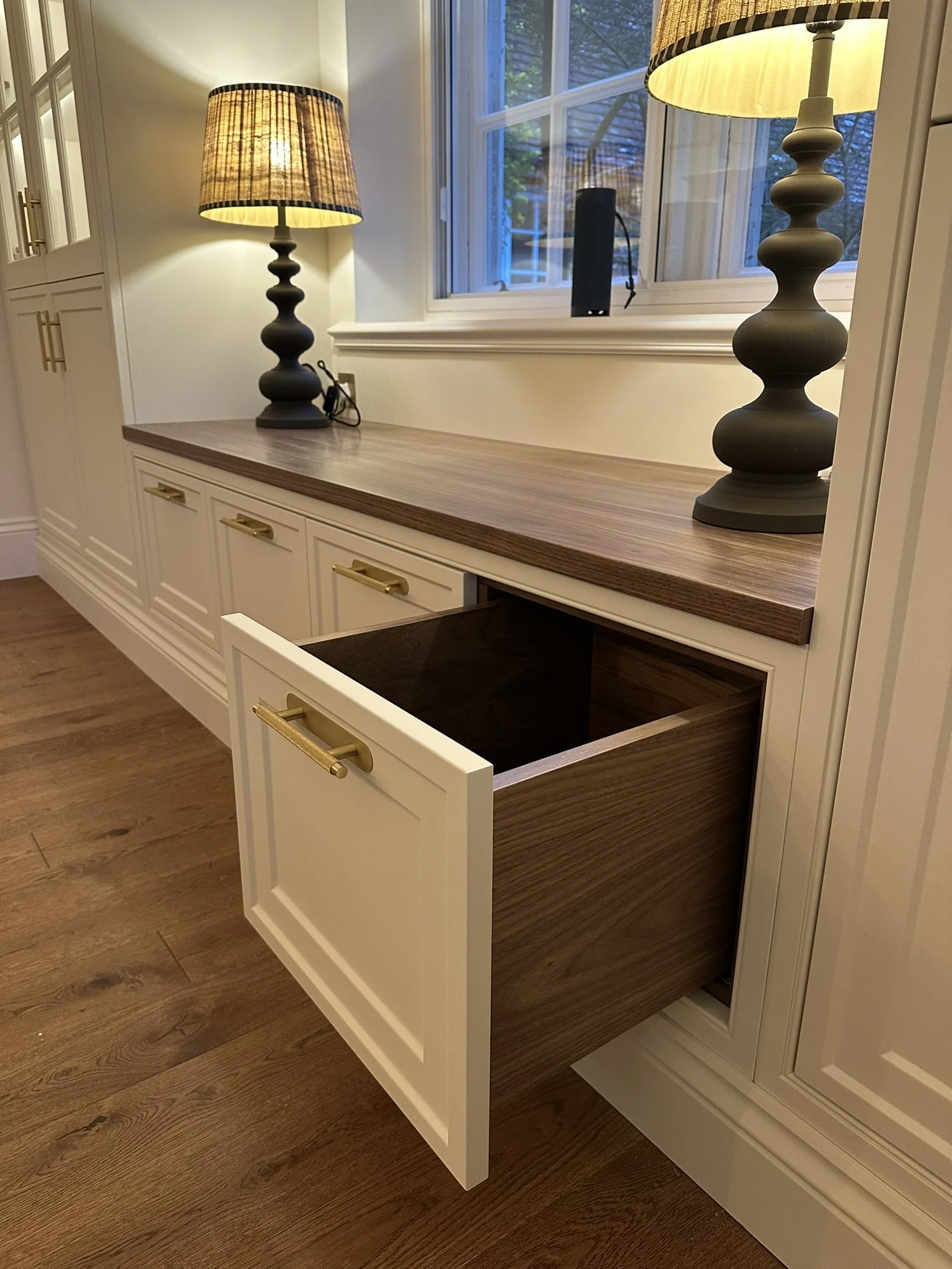 Open drawer in cream-colored cabinet with gold handles, wooden countertop, two black lamps with beige shades on a cream-colored cabinet, window in the background, and hardwood floor.