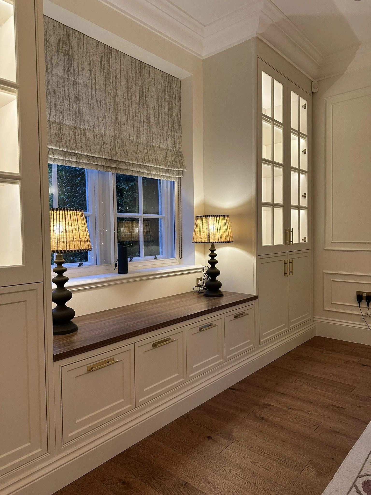 Interior view of a room with a wooden window seat, two matching table lamps, built-in cabinetry with glass-paneled doors, and wooden flooring.
