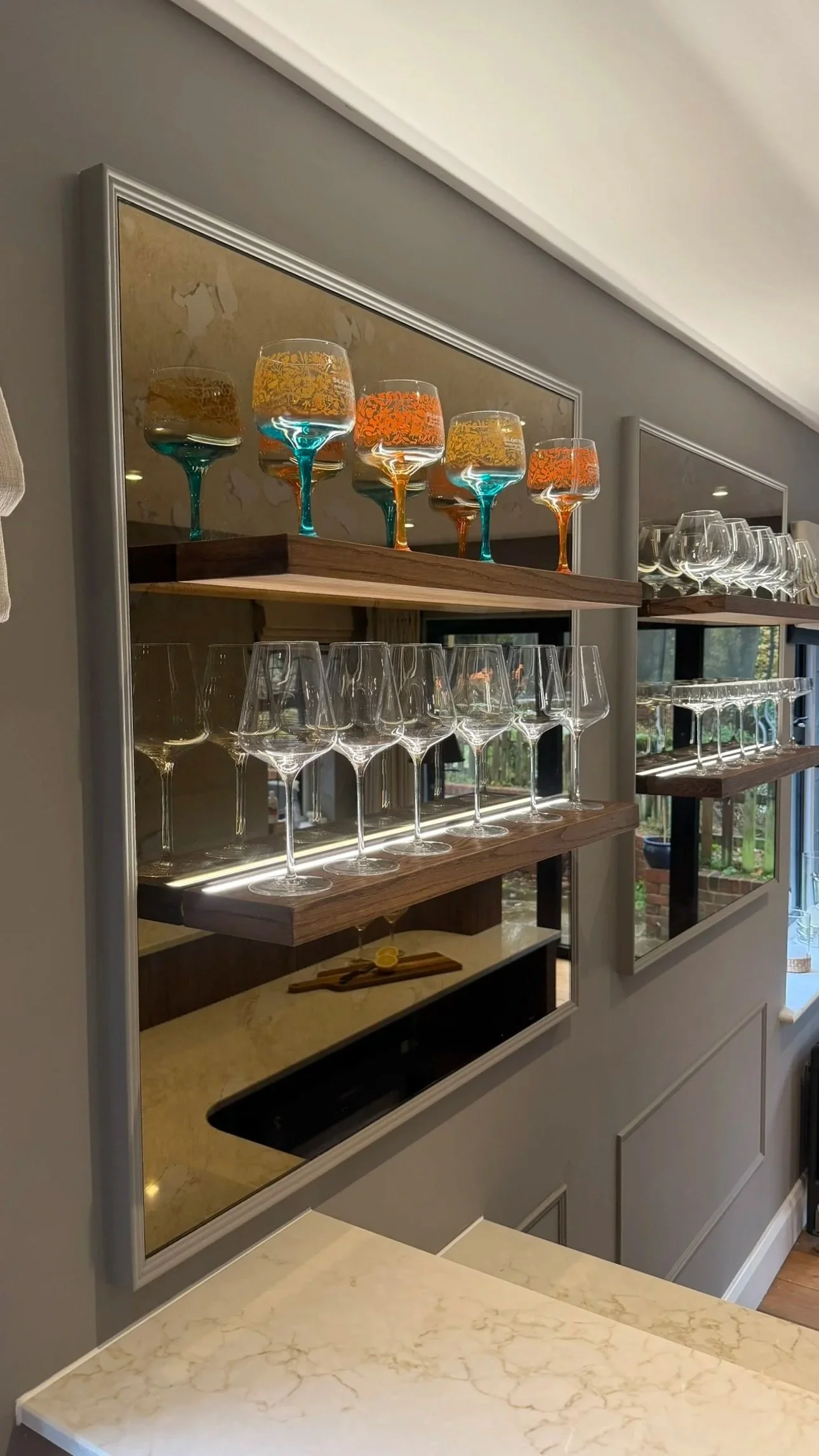Decorative wall shelves with colorful wine glasses and clear tumblers reflected in a mirror, situated above a marble countertop with a lemon and a wooden cutting board.
