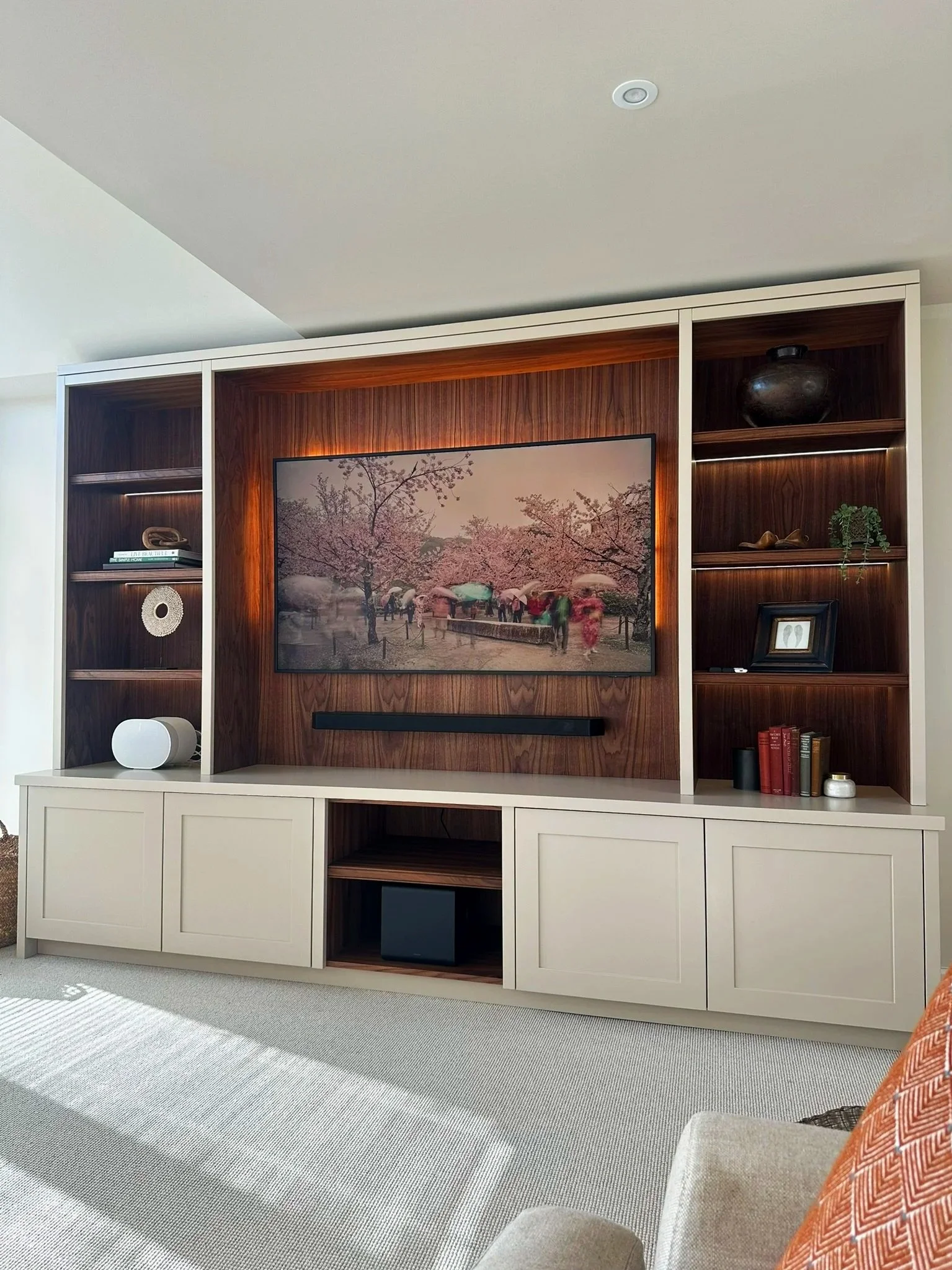 Living room entertainment center with a large flat-screen TV displaying cherry blossom trees, surrounded by open shelves with decorative objects and books, and a beige carpeted floor.