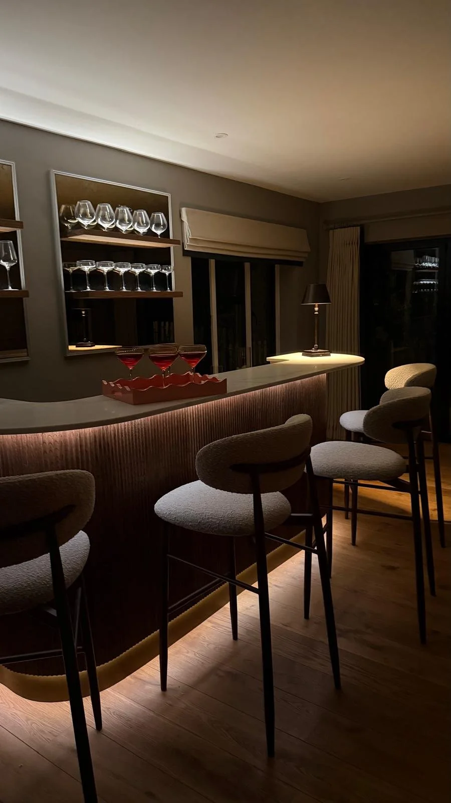 A modern home bar with a curved countertop illuminated by soft ambient lighting. There are four upholstered bar stools in front and shelves in the background with wine glasses and other glassware. The room has neutral-colored walls, wooden flooring, 