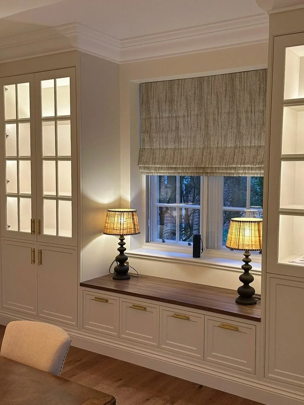 A cozy room with built-in white cabinets, a wooden window seat with two table lamps, and a window with a beige Roman shade.