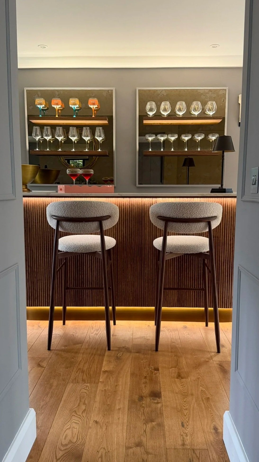 A modern bar with a wooden counter and textured front panel, two white upholstered bar stools, glassware on shelves behind, and soft lighting.
