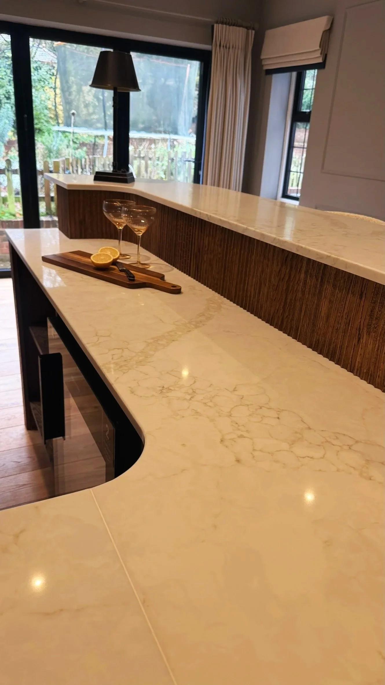 A kitchen island with a marble countertop, a cutting board with lemon halves, two cocktail glasses, and a small knife. In the background, there is a window with curtains, a sliding glass door, and a view of a backyard with a trampoline.