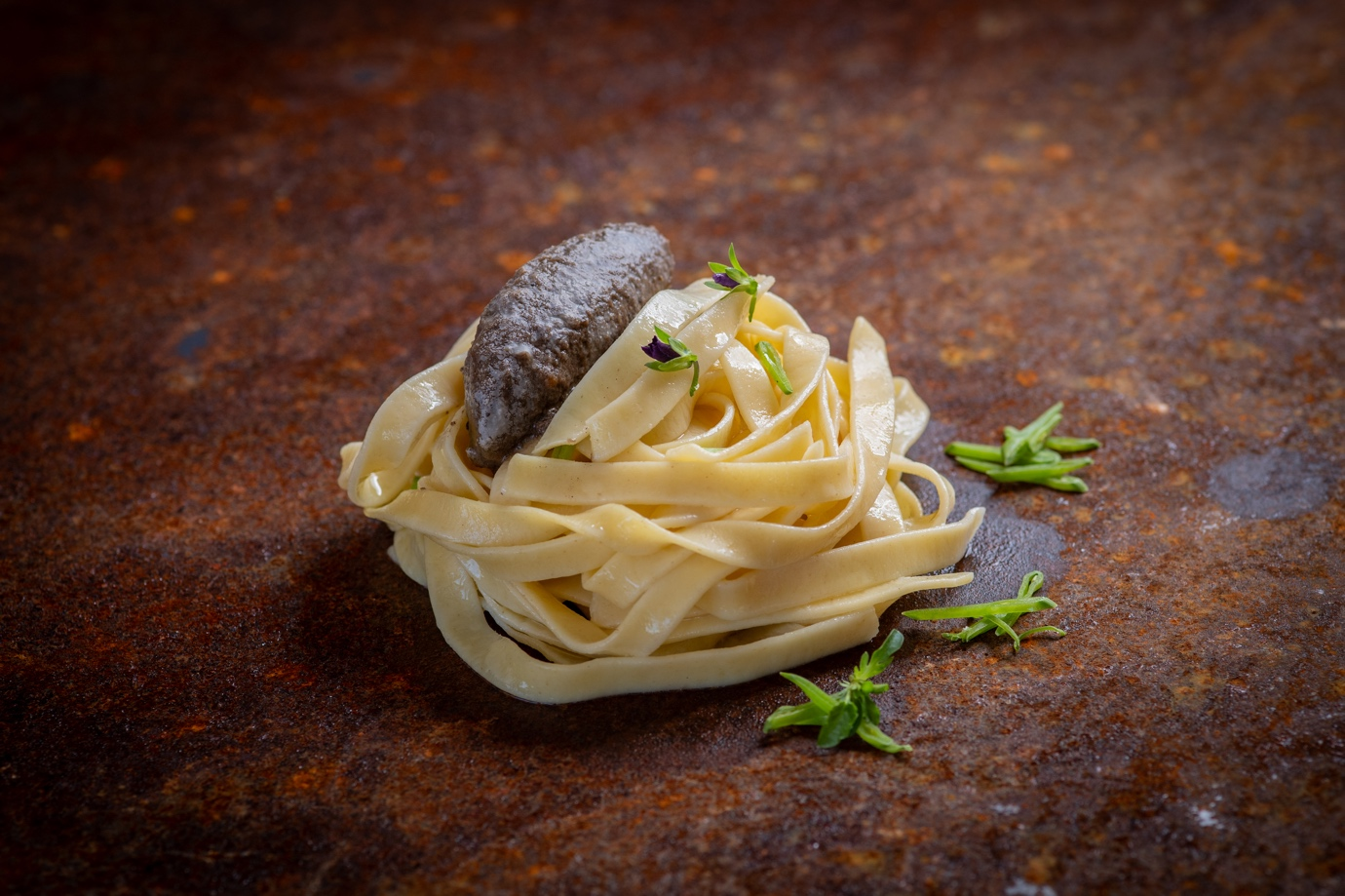 A serving of fettuccine pasta with a dark sausage on top, garnished with green herbs, placed on a rusty surface.