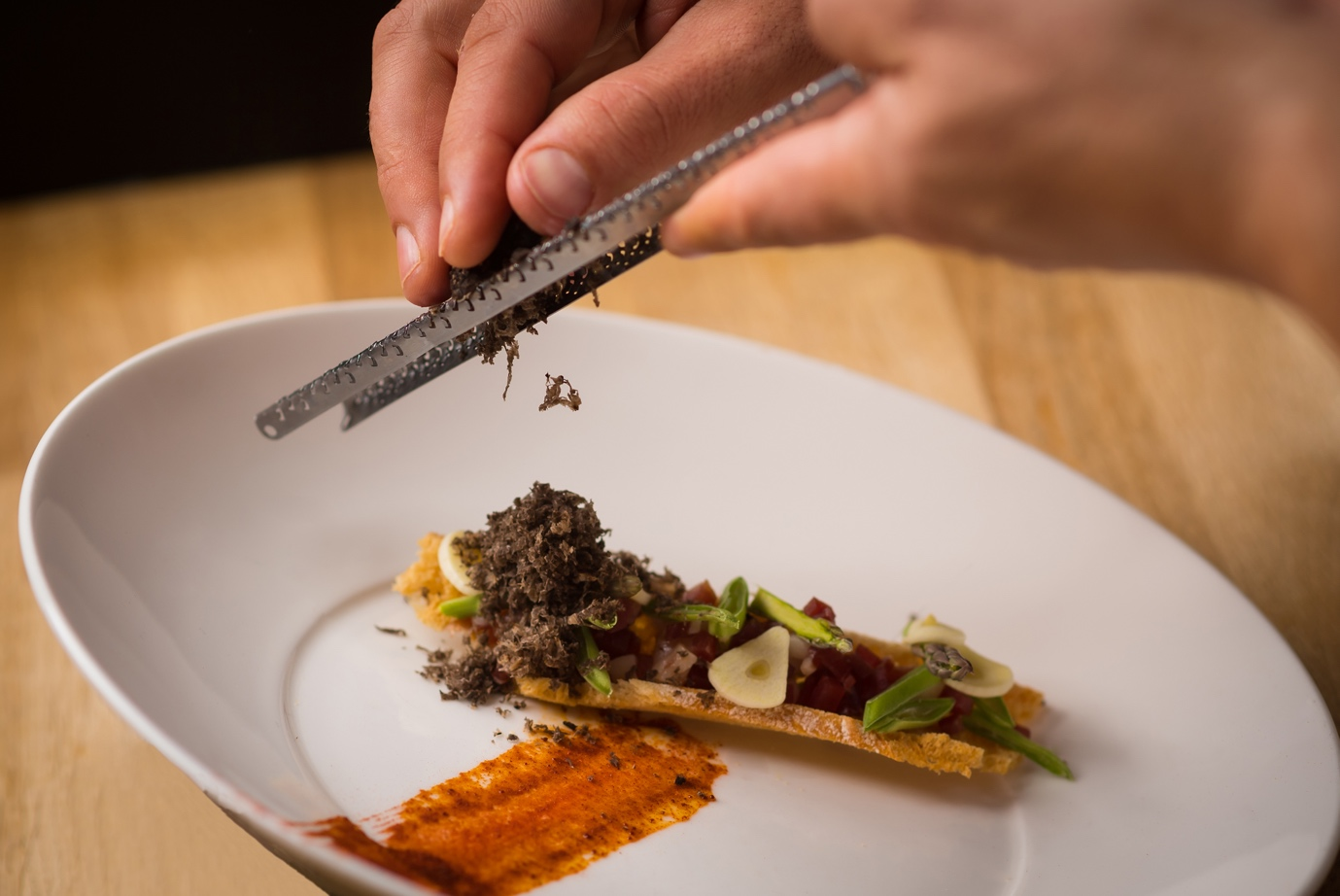 A chef grating black truffle onto a gourmet dish on a white plate, which includes thinly sliced vegetables, herbs, and a crispy base, with a red sauce smear on the side.