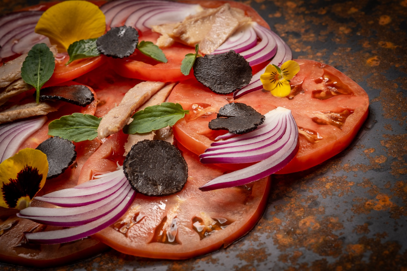 Tomato slices topped with sliced red onions, herbs, black truffle slices, and garnished with edible flowers on a dark, rustic surface.