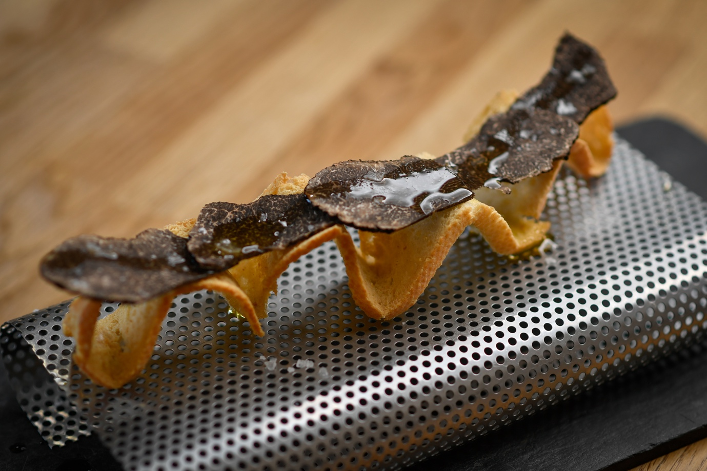Dessert with a crispy shell topped with chocolate and caramel sauce on a perforated metal tray.