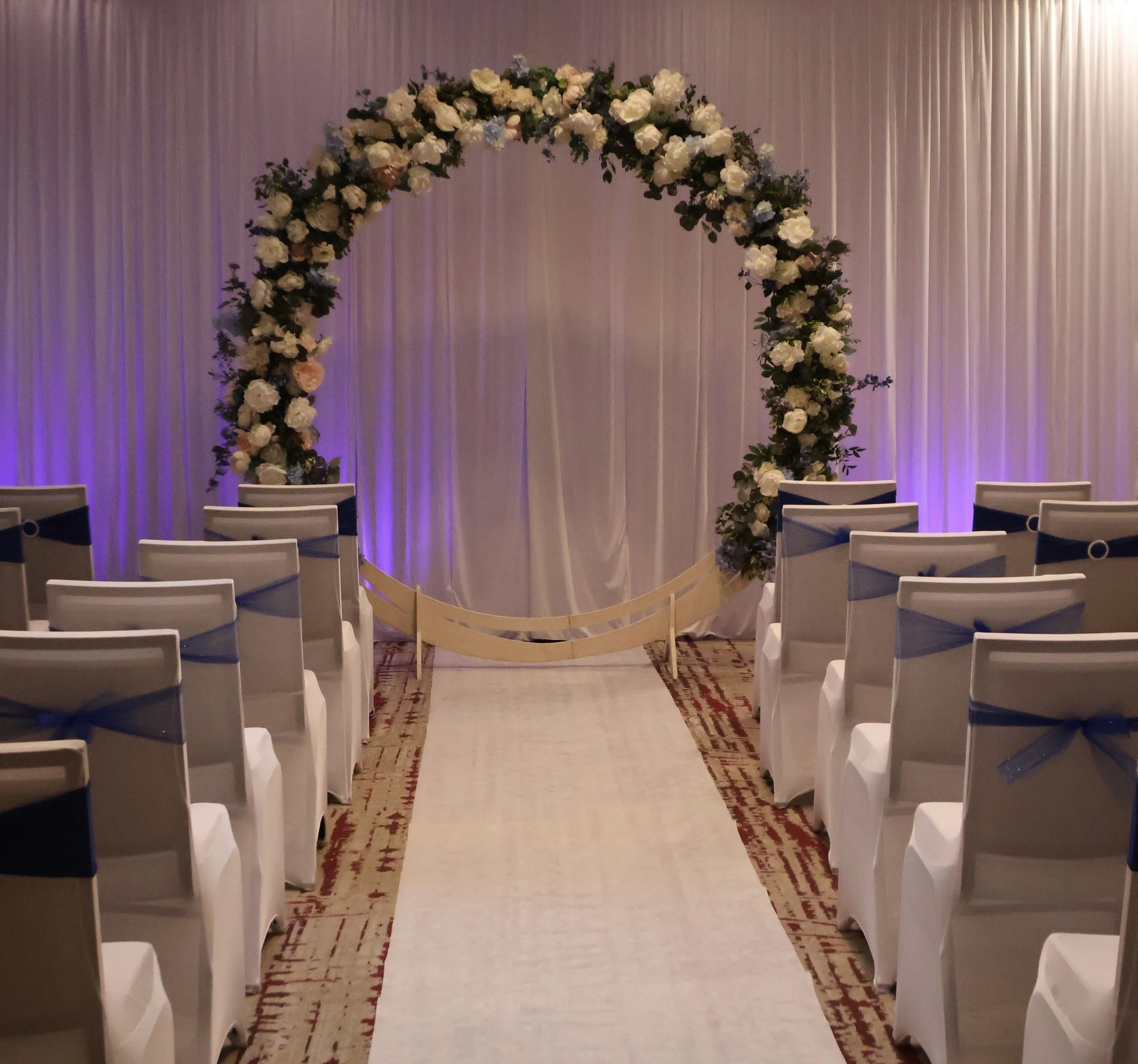 Indoor wedding ceremony setup with white chairs decorated with navy blue sashes, facing a floral arch over an aisle, with purple lighting and draped curtains in the background.