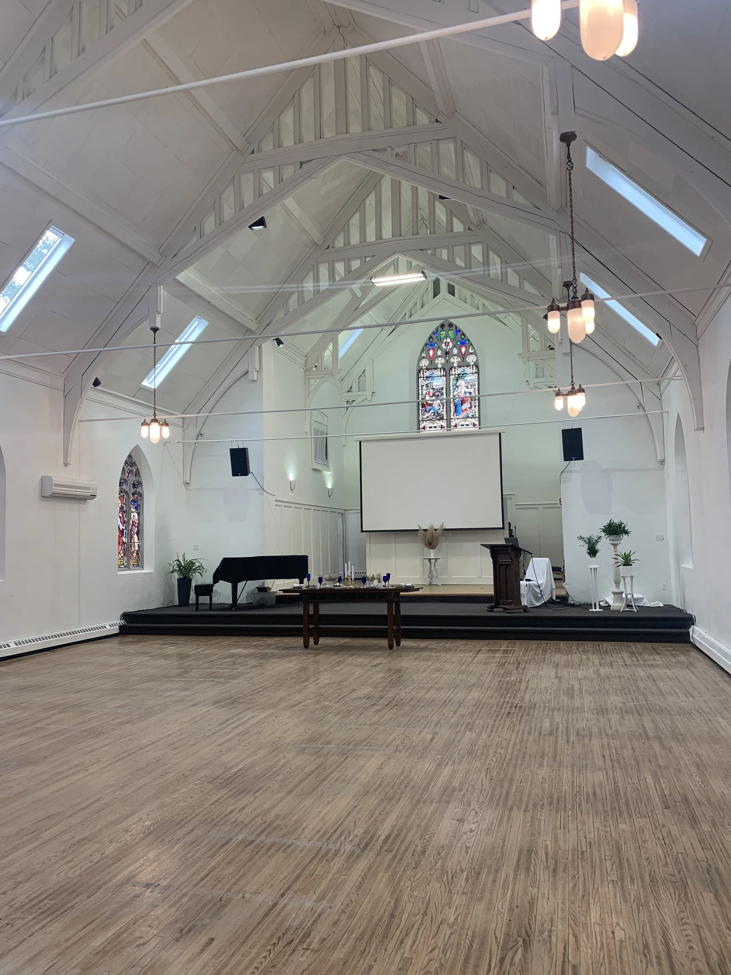Church interior with high vaulted ceiling, stained glass windows, wooden floor, stage with piano and lectern, and modern lighting.