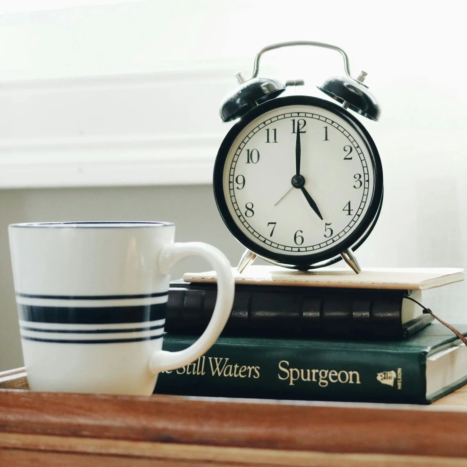A black and white alarm clock showing 4:00, placed on a closed book titled "Still Waters" and "Spurgeon." A white and black striped coffee mug is in the foreground, and a white wall is in the background.