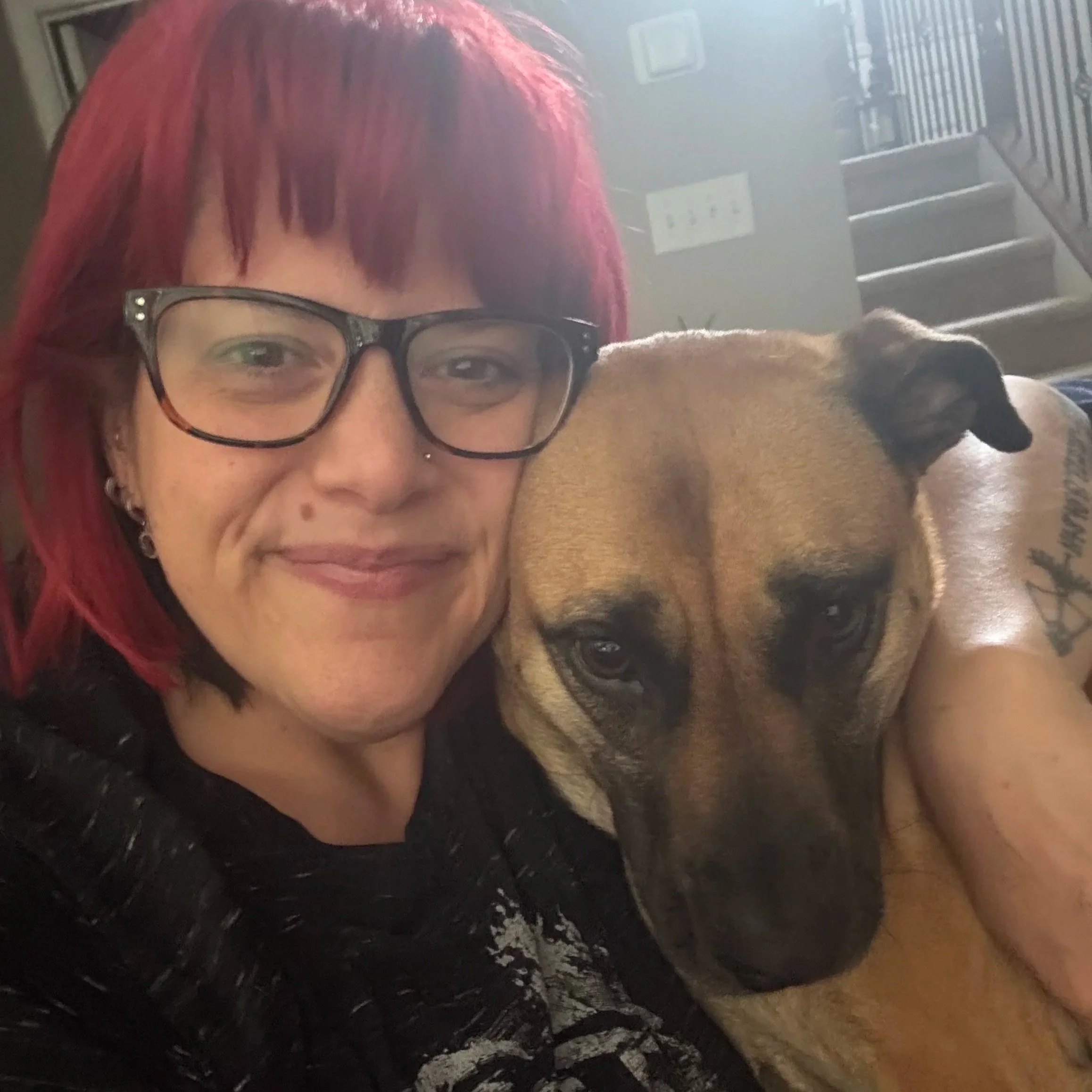 A woman with red hair and glasses smiling with a dog, possibly a boxer, resting its head on her shoulder.
