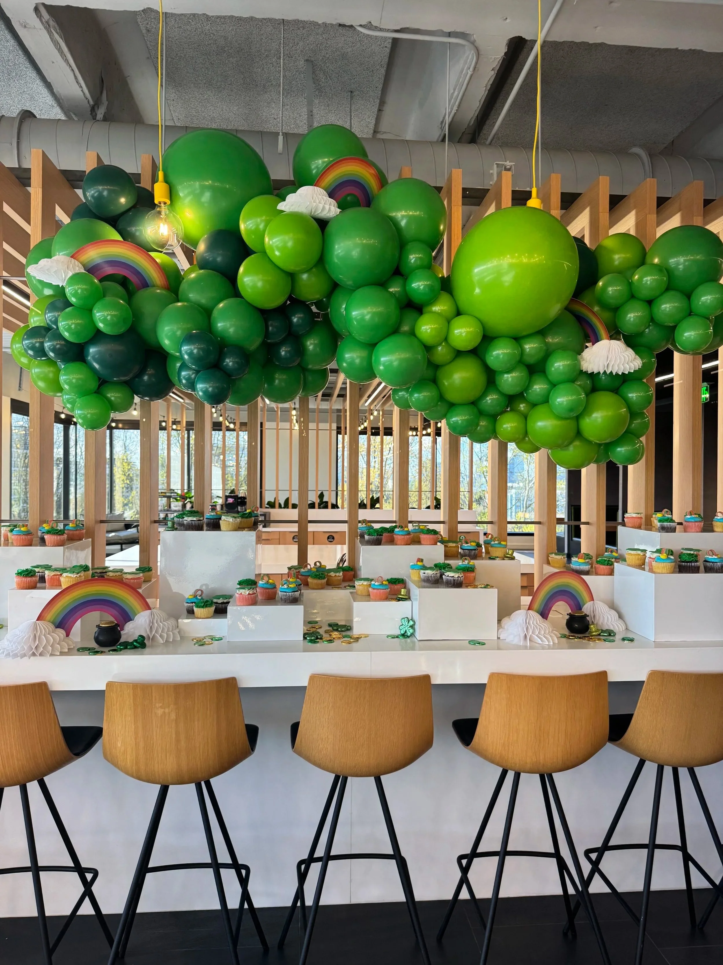 Indoor party setup with colorful balloon decorations, rainbow and cloud cutouts, cupcake display, and wooden chairs