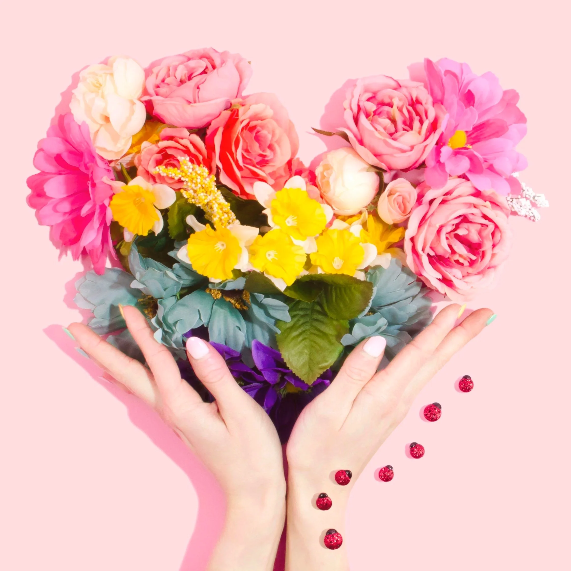 Two hands hold a colorful bouquet of pink, white, and yellow flowers against a pink background, with red and black jeweled pins scattered on the surface below.