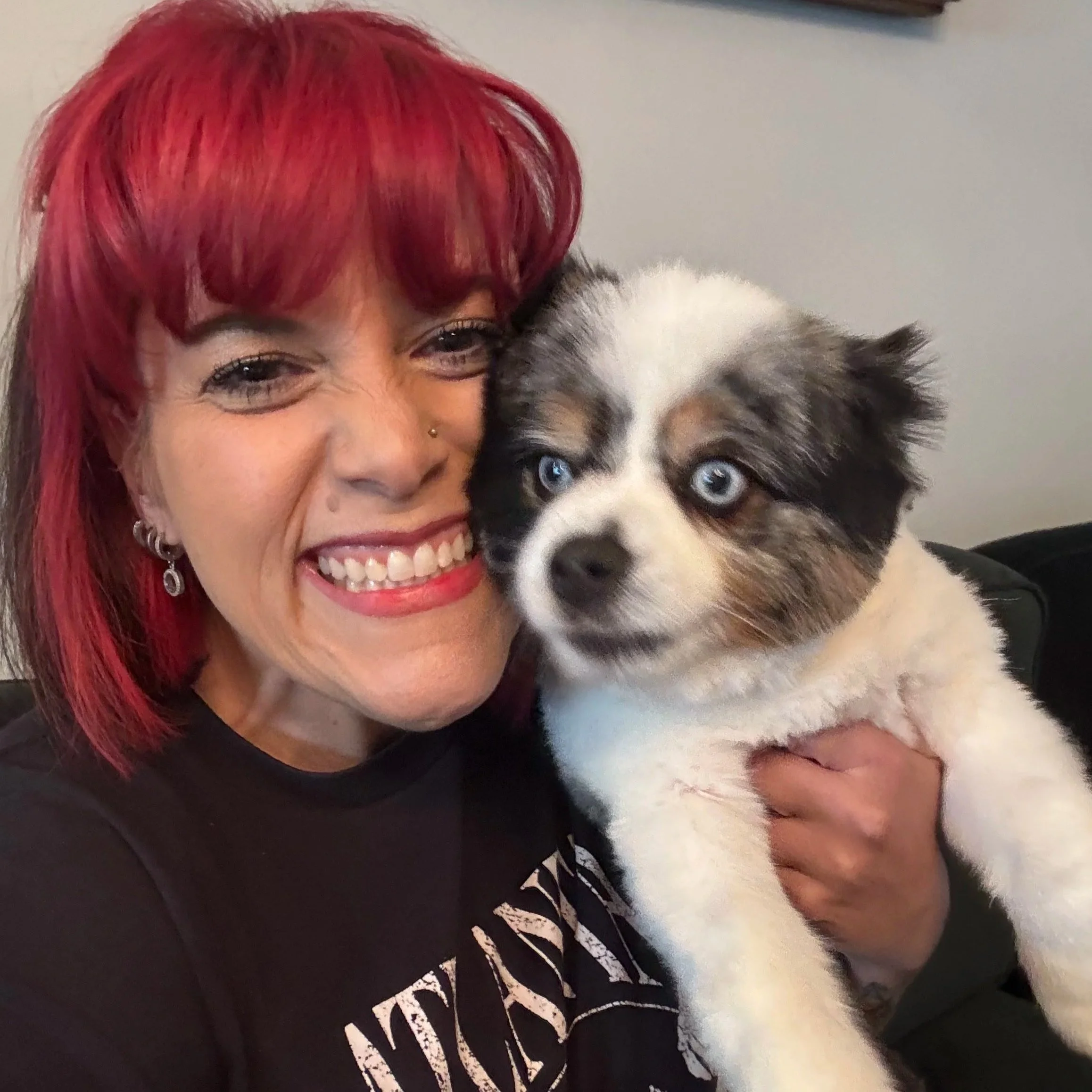 A woman with bright red hair smiling, wearing earrings, with a dog that has blue eyes and a black, white, and gray fur coat close to her face.