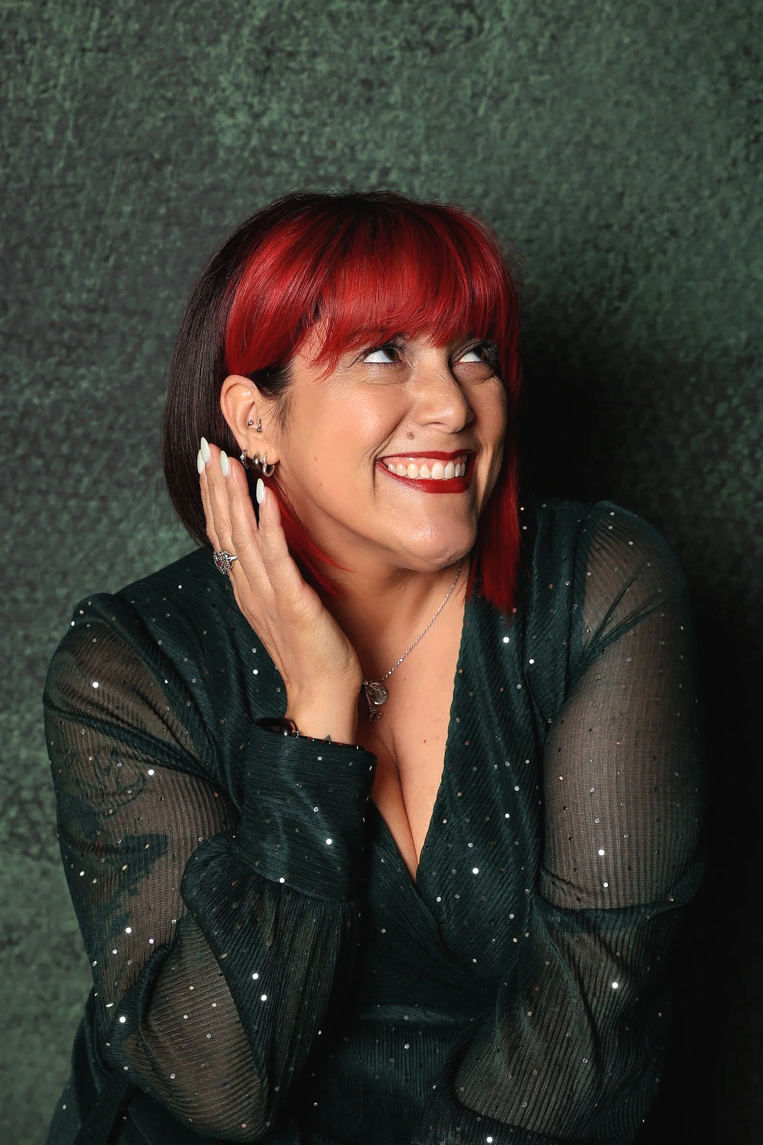 A woman with red and black hair smiling, wearing a black sheer top with small white dots, and silver jewelry, against a textured dark green background.