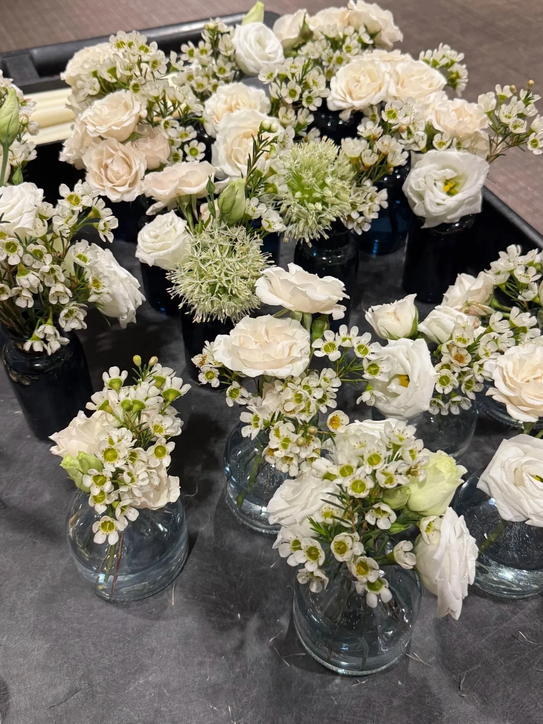 Multiple small glass vases filled with white flowers, including roses and daisies, arranged on a dark surface.