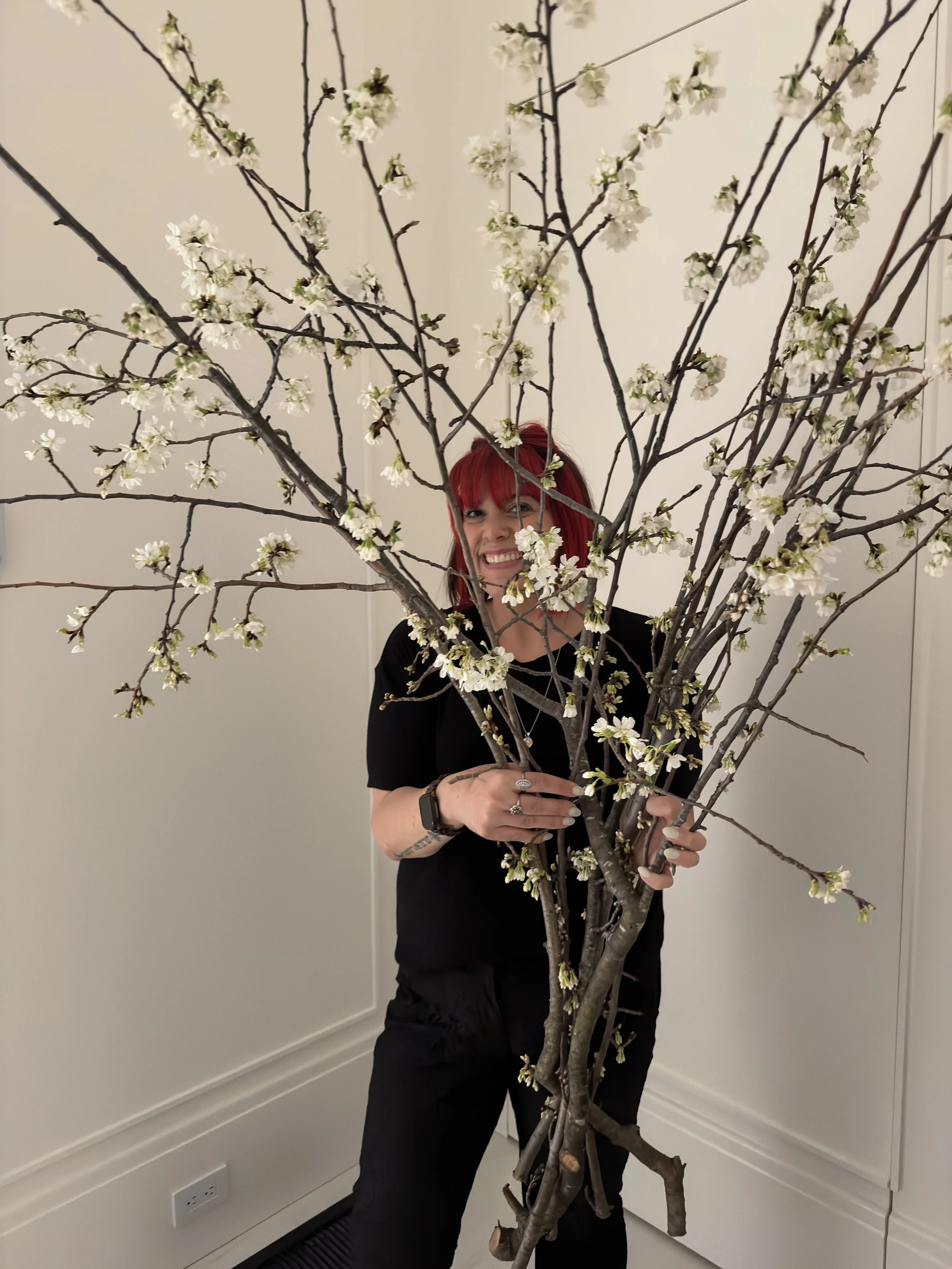 A person with red hair standing behind a large, flowering tree branch with white blossoms, smiling at the camera, indoors with plain white walls.