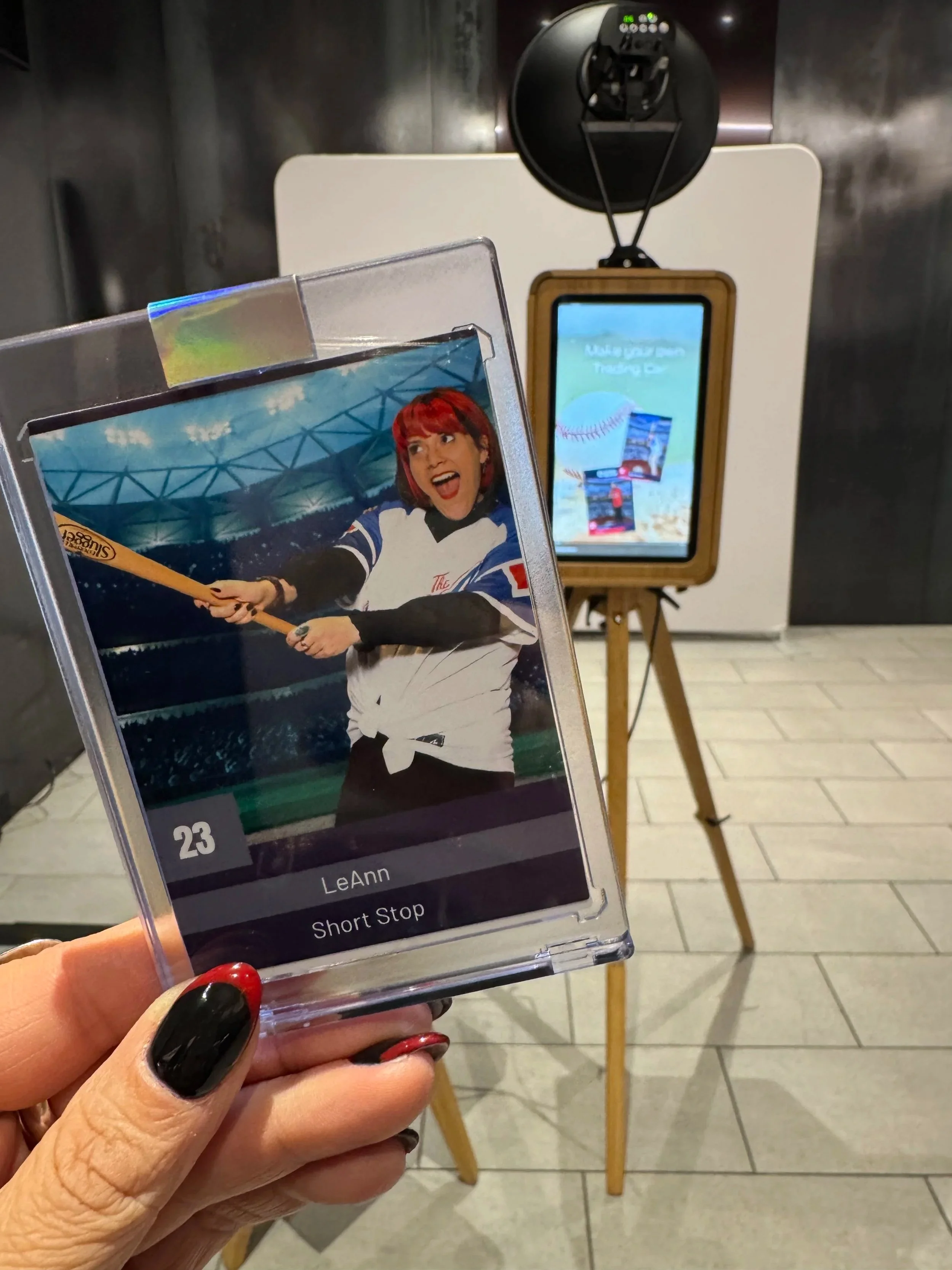 Close-up of a person holding a laminated card with a picture of a woman in a sports jersey with red hair, holding a baseball bat and smiling. In the background, there is a signboard with a baseball graphic and some images, along with a black smoke detector mounted on a wooden tripod.