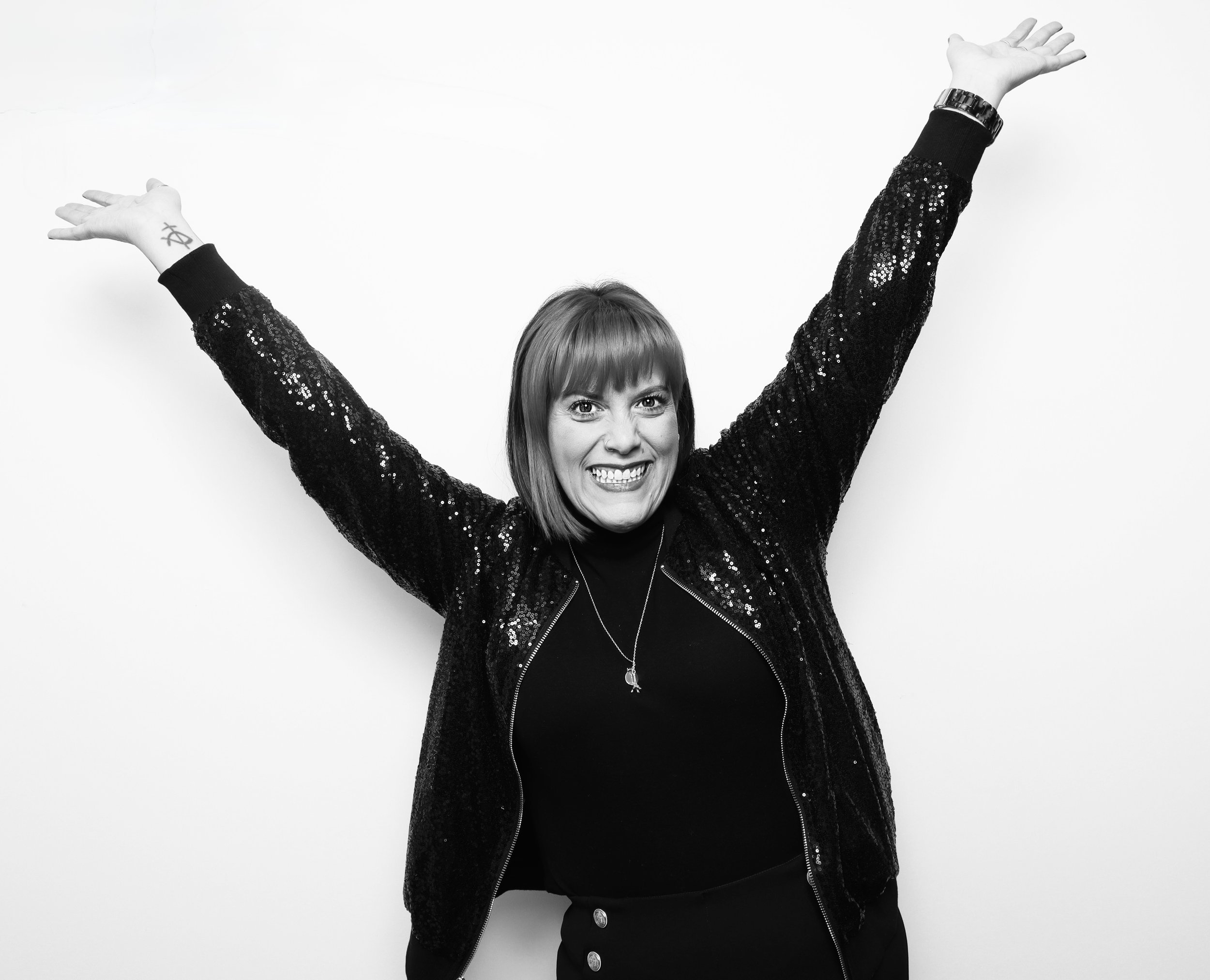 A woman with shoulder-length hair, wearing a black sequin jacket and a black top, smiling with arms raised against a plain white background.