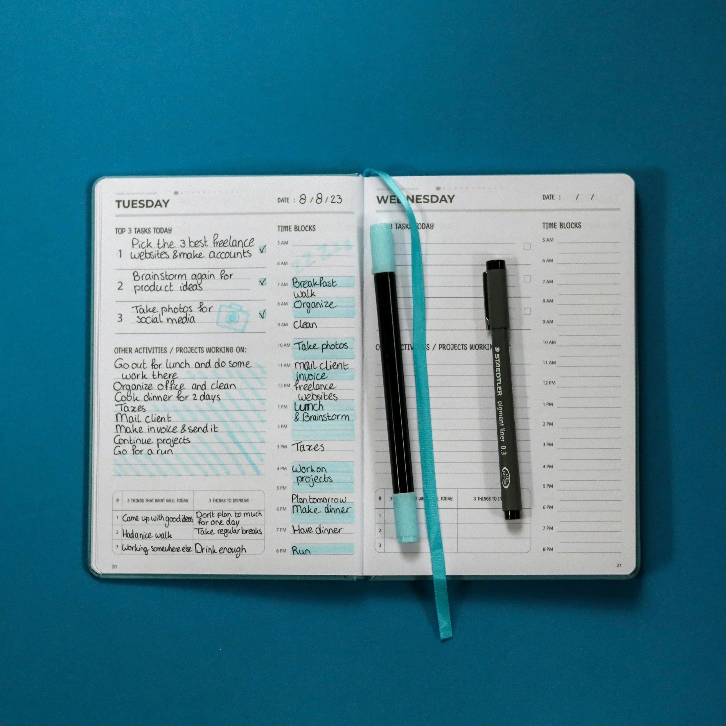 Open planner on a blue surface, showing weekly schedule with handwritten tasks and blue sticky notes for task details, with two black pens and a turquoise pen placed on the planner.