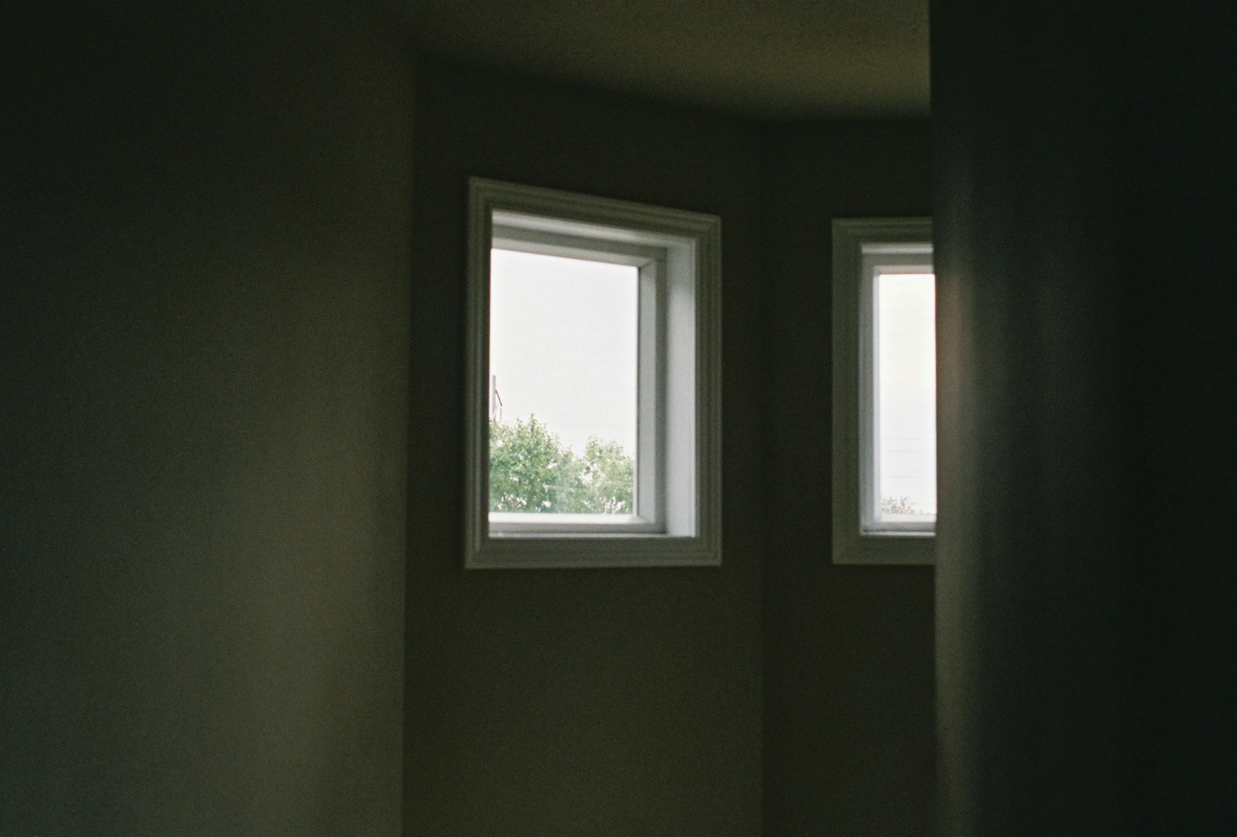 Two small square windows on a plain wall let in soft natural light, with faint greenery visible outside. The room is dimly lit, creating a quiet and minimal atmosphere.
