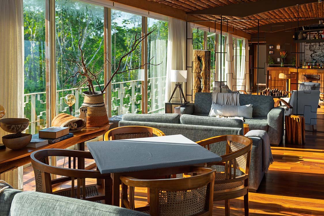 Bright living room with large windows showing green trees outside, sunlight streaming in, a dark gray sofa with pillows, a wooden dining table with chairs, decorative vases, and modern interior design elements.
