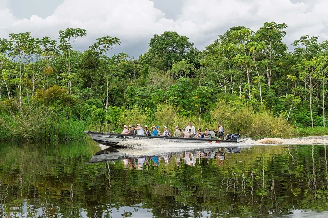 A group of people on a boat moving on a calm river with lush green trees along the riverbank and a cloudy sky overhead.