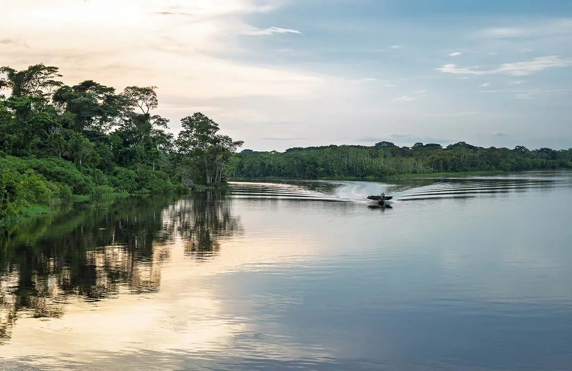 A serene river scene with calm water reflecting green trees along the riverbank under a cloudy sky, and a small boat speeding across the water creating ripples.
