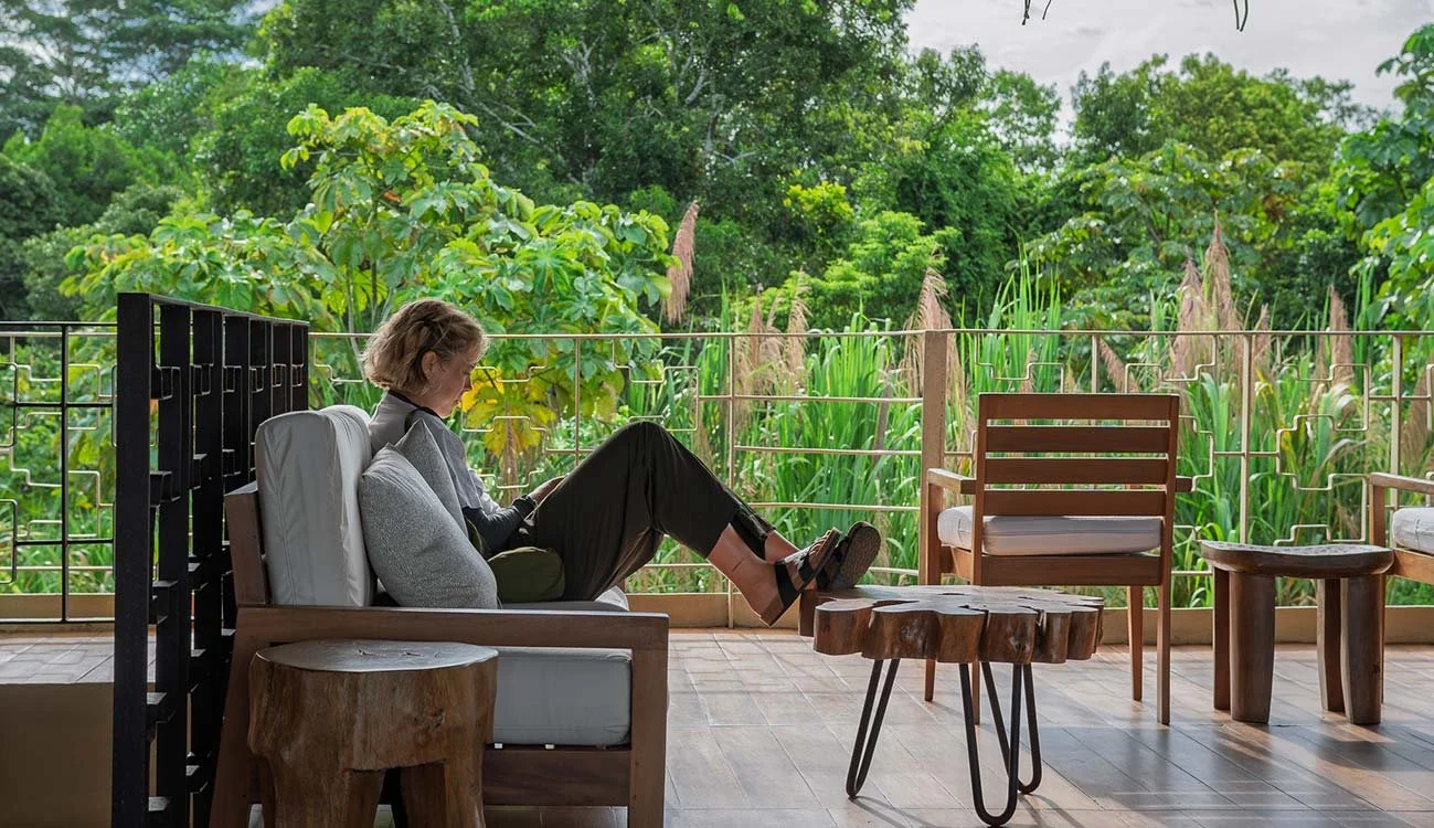 A woman sitting on a sofa on a balcony, reading a book, surrounded by lush green trees and plants.