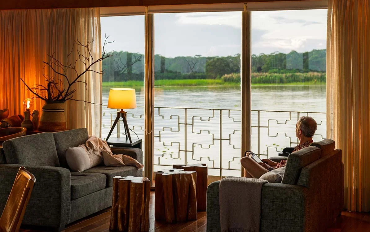 A cozy living room with large window overlooking a river, a gray sofa with pillows, a side table with a lamp, a woman sitting in a chair reading, and decorative tree trunk tables.