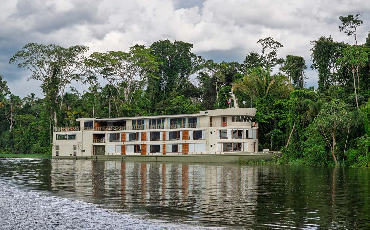 Large white multi-story houseboat with multiple windows and outdoor decks on a river next to lush green forest and cloudy sky.