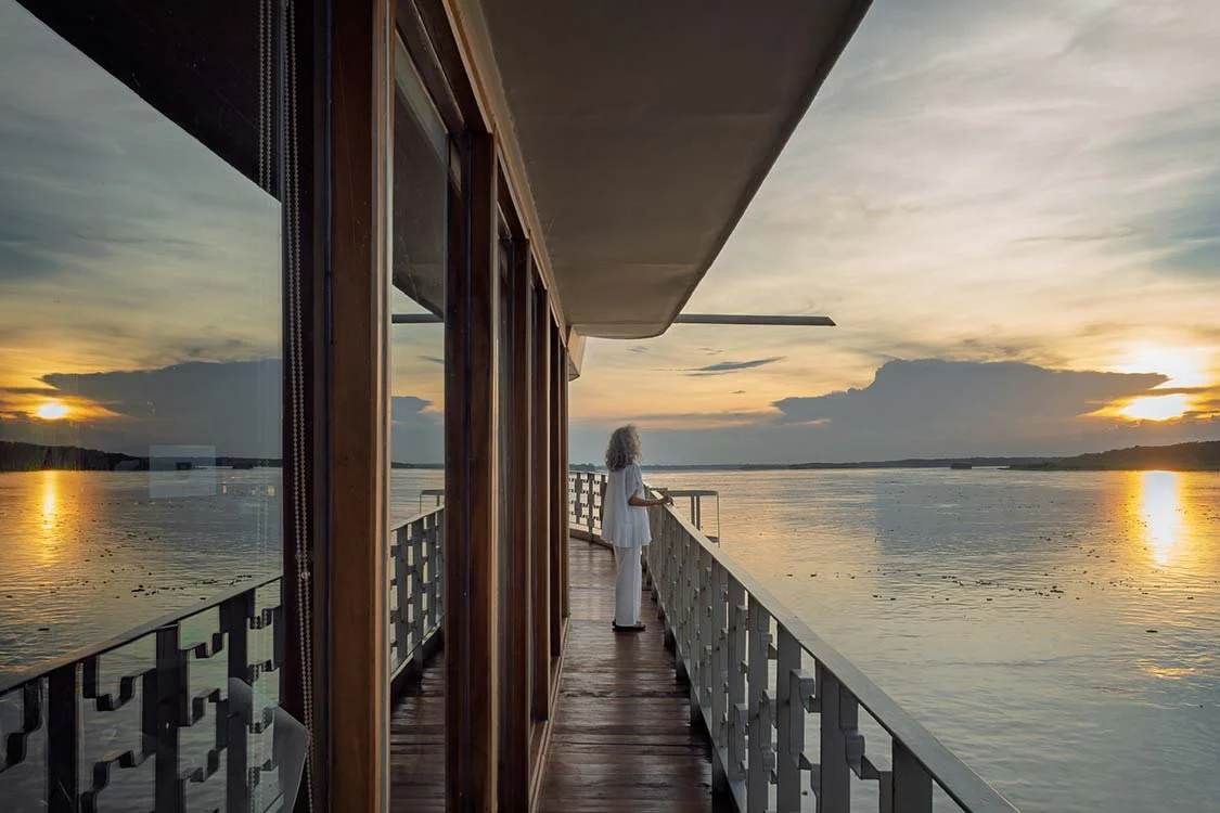 A woman dressed in white standing on a balcony overlooking a body of water during sunset.