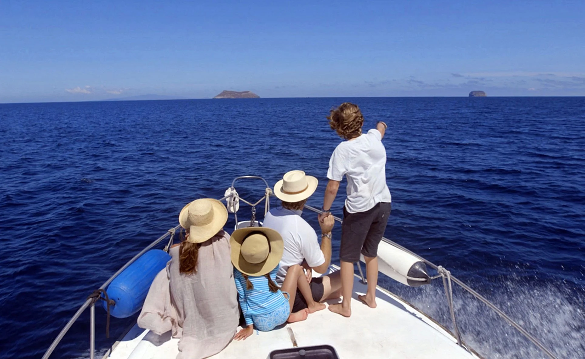 A group of people on a boat in the ocean, with some pointing towards the distant islands on the horizon, wearing summer clothing and hats, enjoying a sunny day.