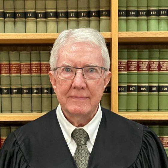An elderly man with gray hair and glasses wearing a black robe and a gray tie, standing in front of a bookshelf filled with law books.