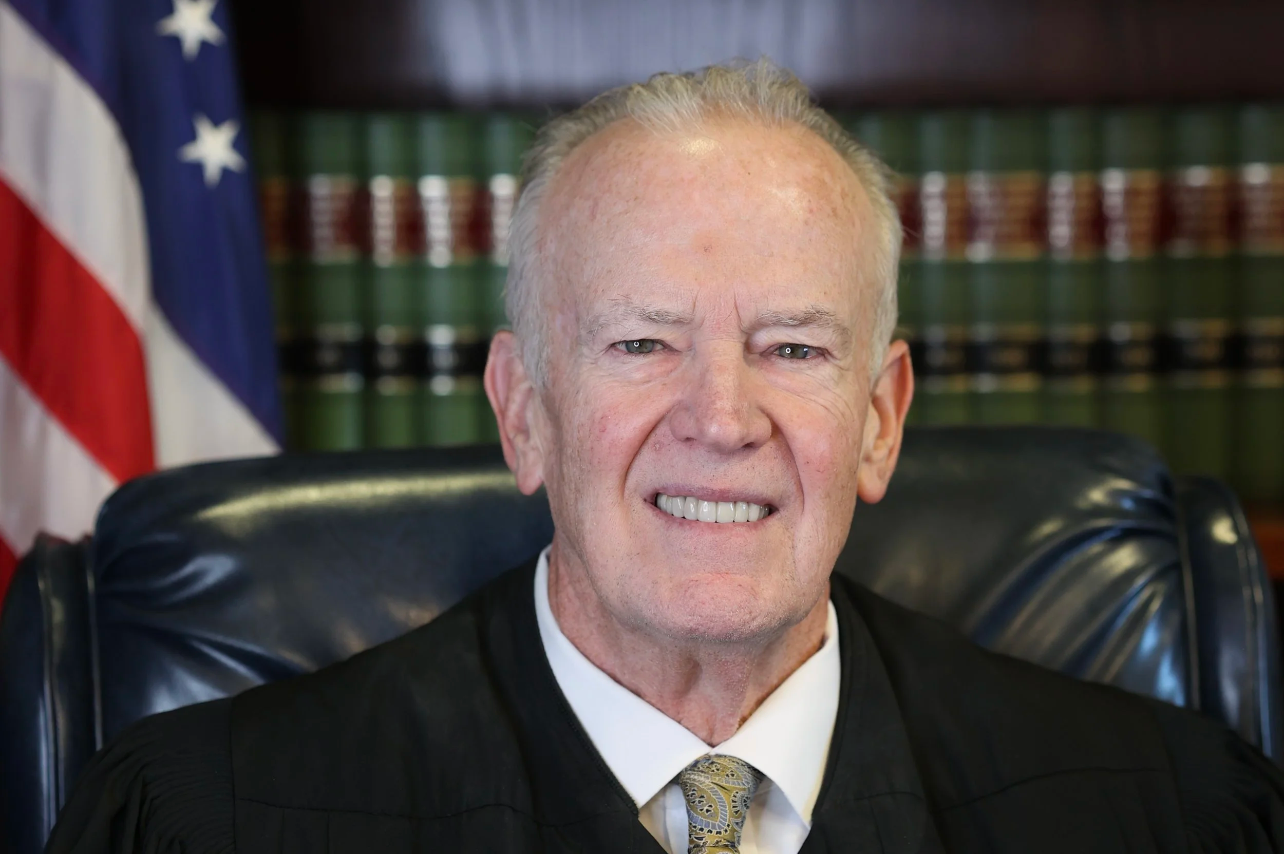 A judge in black robes sitting in a courtroom with an American flag and bookshelves with legal books in the background.