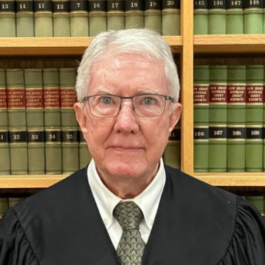 A woman with short gray hair and glasses wearing a black robe over a white shirt with a patterned tie, standing in front of a bookshelf filled with legal books.