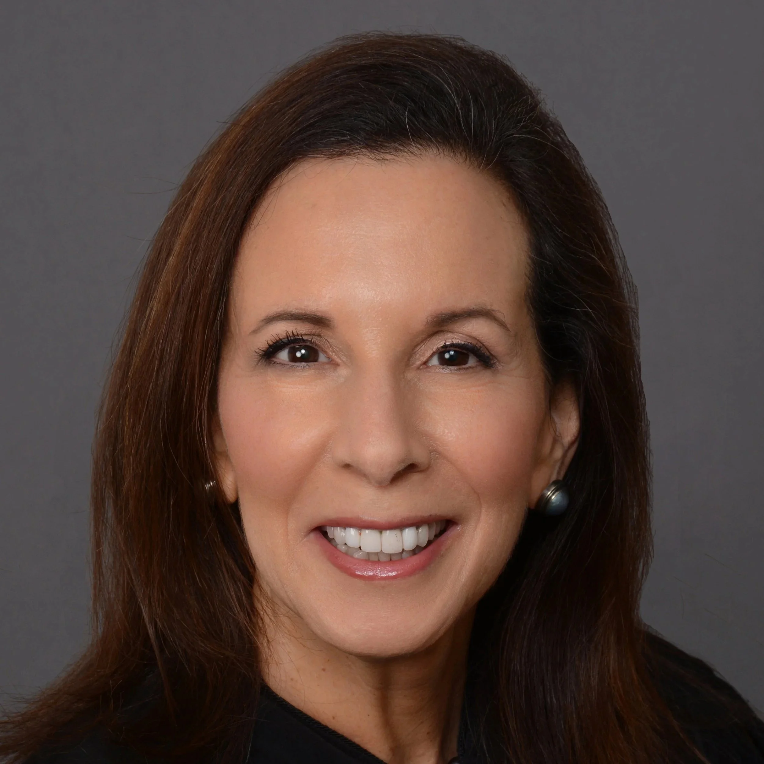 A judge sitting in a courtroom, wearing a black robe, with a bookshelf of legal books behind him and a partial view of an American flag on the left.