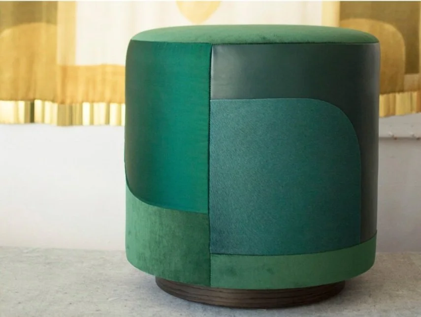 An emerald green Decoupage upholstered cylindrical stool with a wooden base, positioned on a white surface in front of yellow curtains.