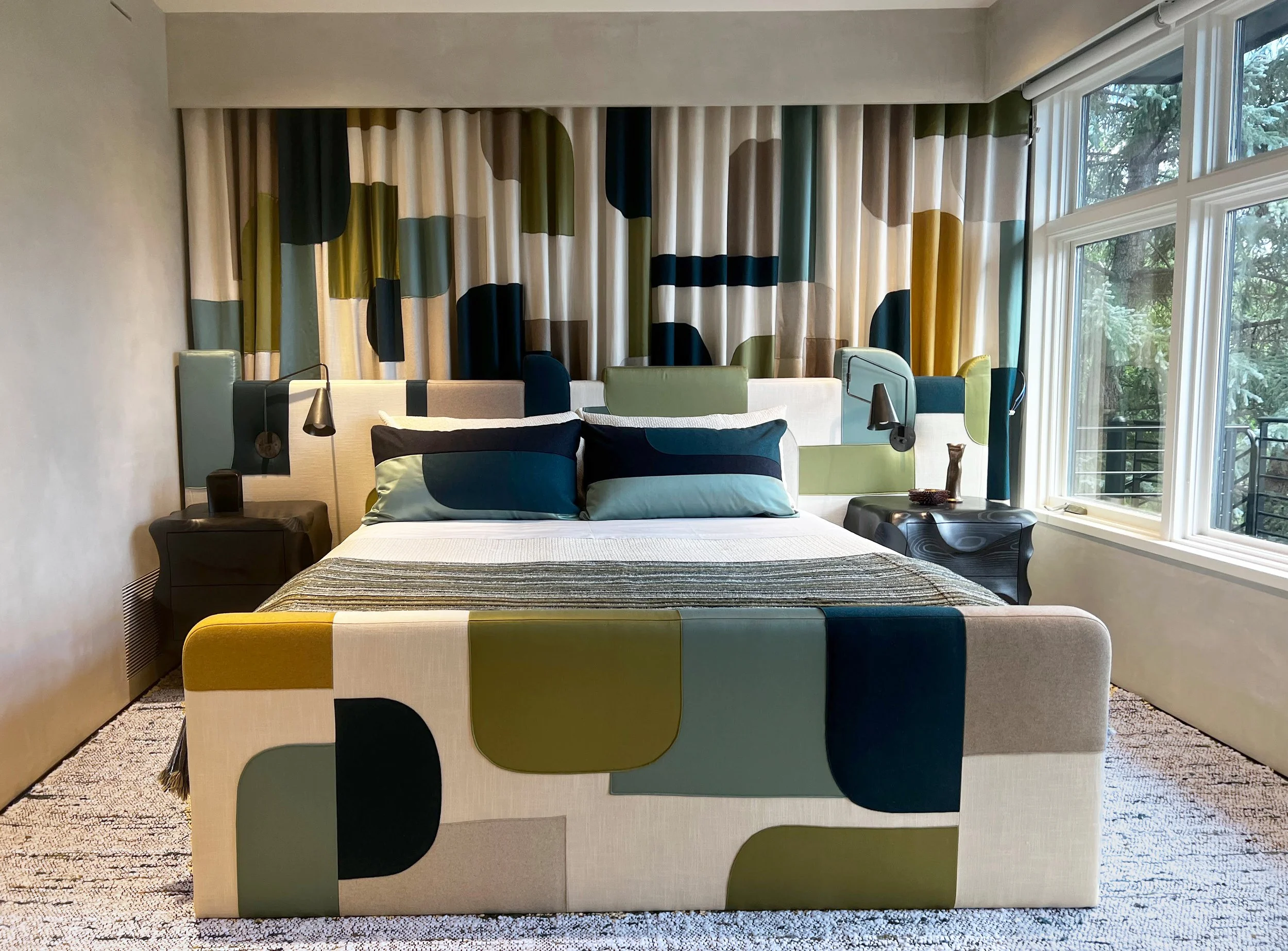 A Decoupage bedroom with a colorful, geometric-patterned headboard and matching curtains, two black side tables with lamps, a bed with striped blankets, two pillows, and large windows with a view of trees outside.