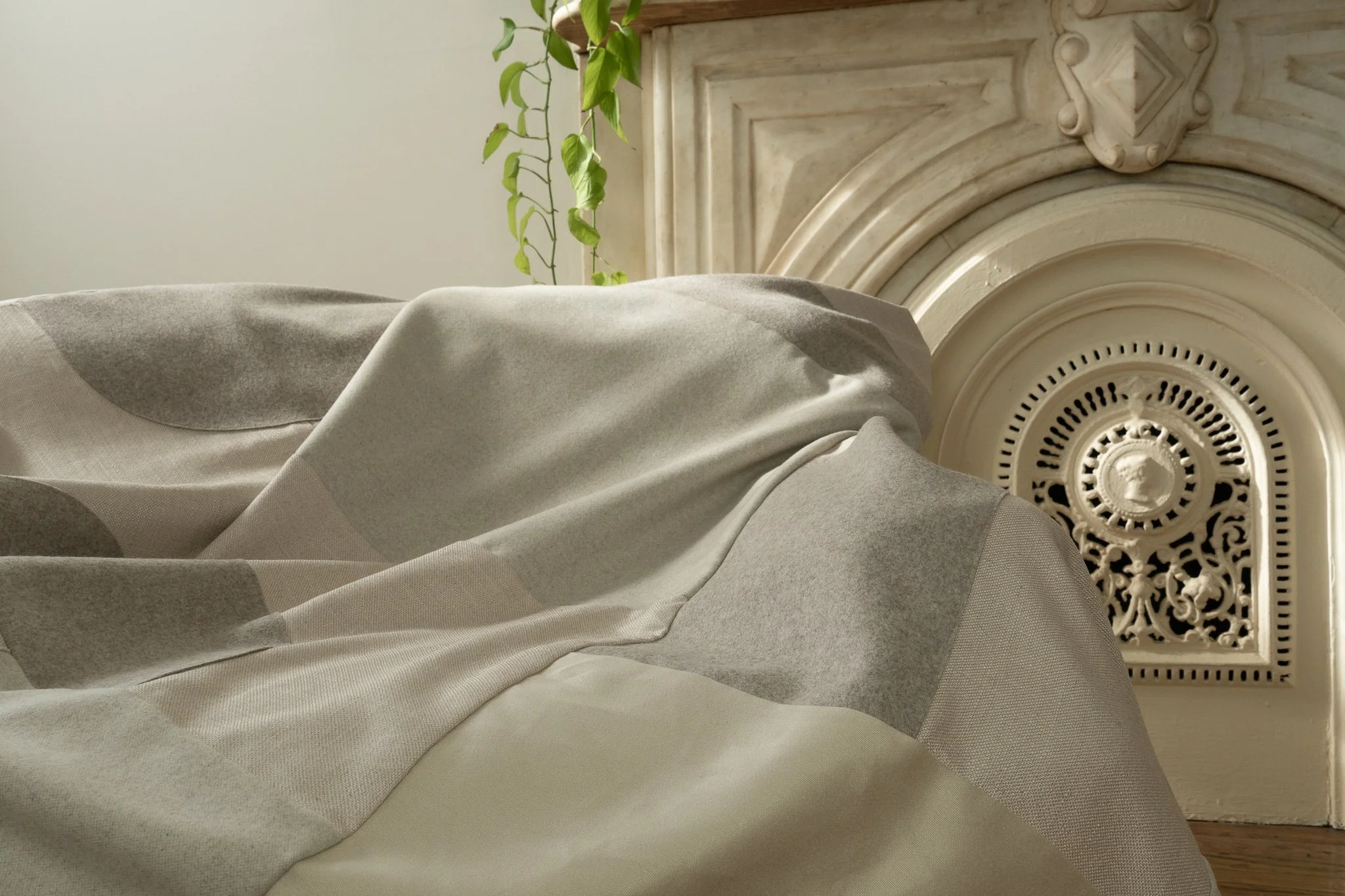 Close-up of a bed with a grey and beige patterned blanket, next to a vintage style fireplace with ornate details and a small green plant hanging over it.