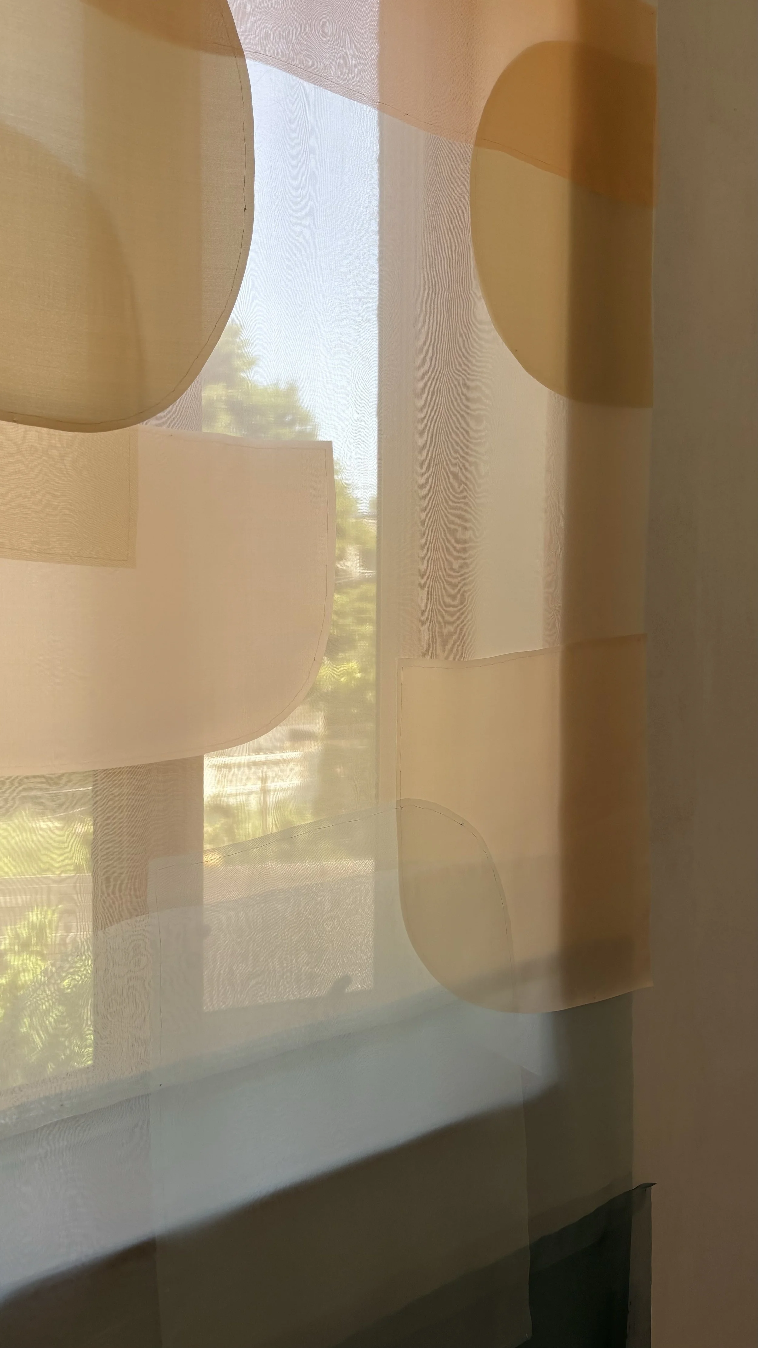 Glass window with Decoupage drapery of peach and amber-colored geometric shapes, sunlight filtering through, trees visible outside.