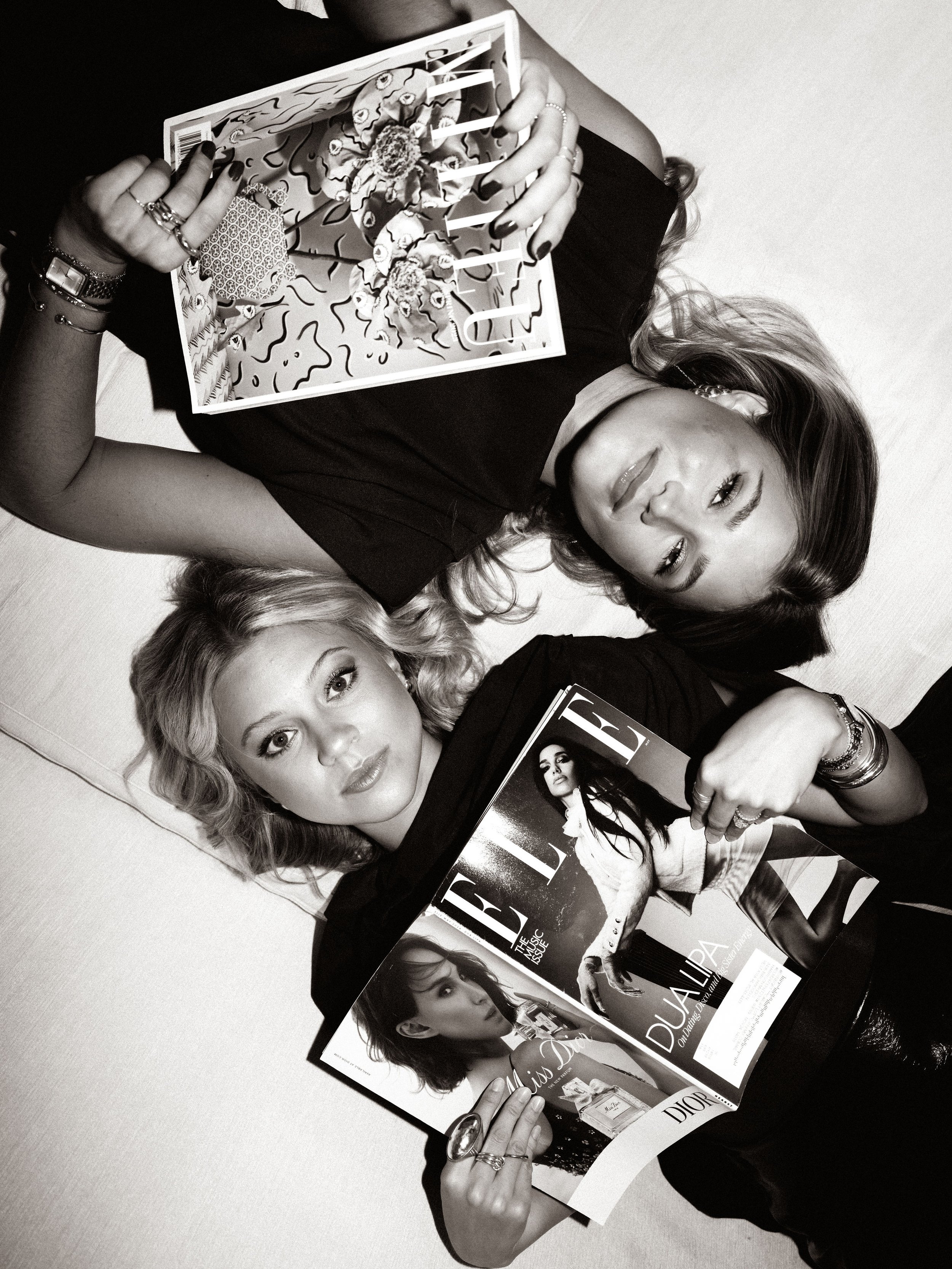 Black and white photo of a woman lying down, holding an issue of Vogue magazine featuring a woman on the cover, with her eyes slightly open and hair styled in waves.