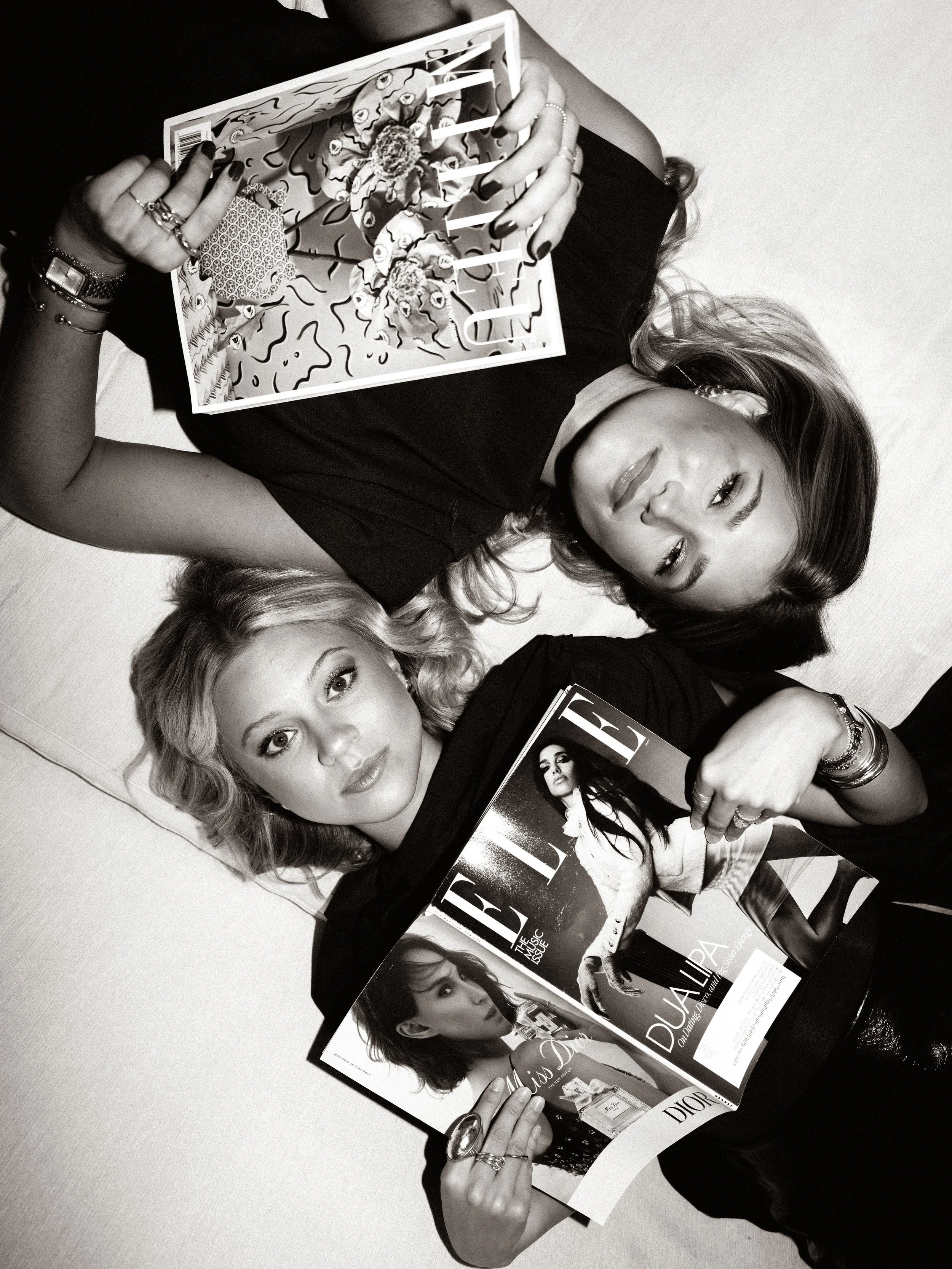 Black and white photo of two women lying on the floor, holding magazines. One magazine features a woman on the cover, and the other magazine is a fashion magazine titled 'Dior.' The women have styled hair and are wearing jewelry, with one woman winking at the camera.