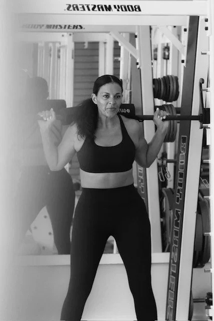 A woman lifting a barbell in a gym, with weights on her shoulders, standing in front of a mirror.