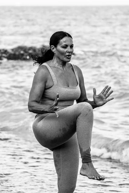 A woman practicing yoga by the water on a beach, balancing on one leg with her arms raised.