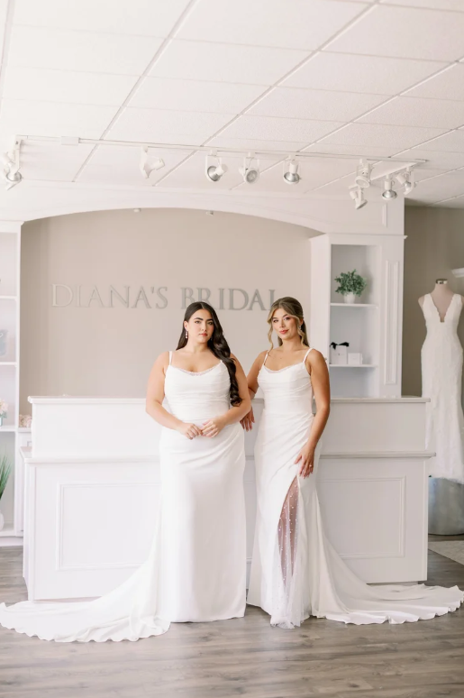 Two women in white wedding dresses standing inside a bridal store with a sign reading "Diana's Bridal" behind them.