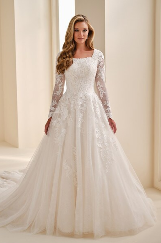 A woman wearing a white wedding dress with lace long sleeves and a full skirt, standing indoors.