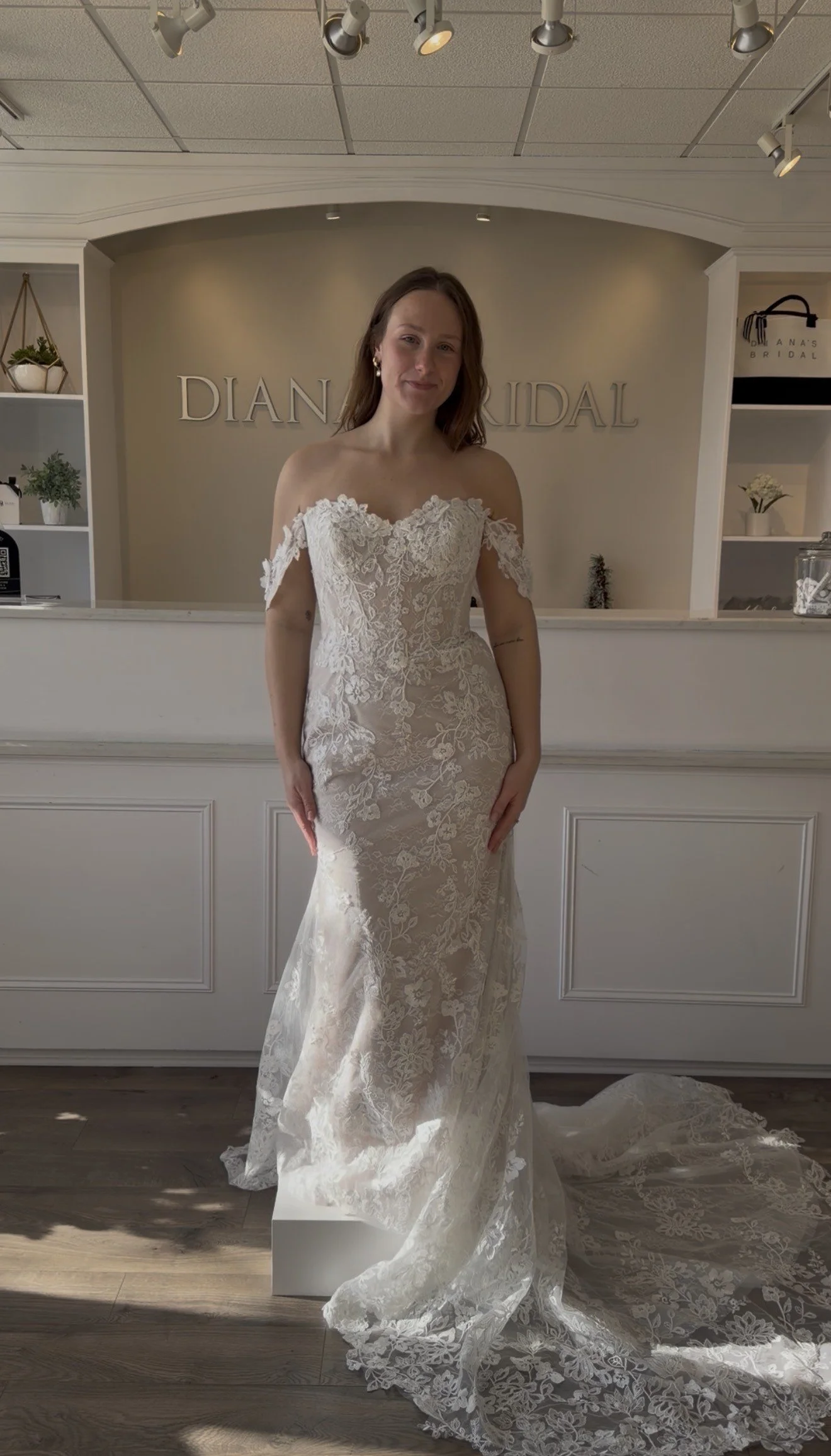 Woman in a lace wedding dress standing in bridal shop. Sign reads 'Diana Bridal' in background.